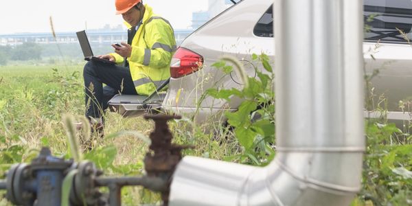 Worker in safety gear using laptop and phone near industrial piping outdoors.