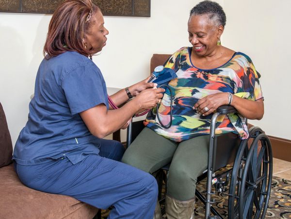 A nurse checks blood pressure of a smiling woman in a wheelchair.
