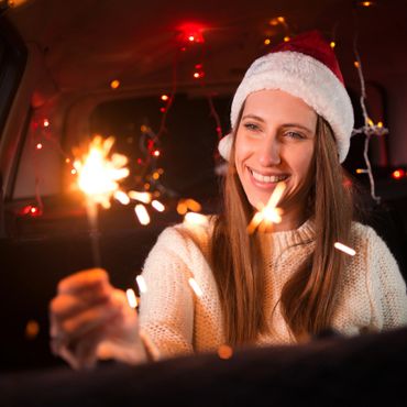Woman in Santa hat holding a sparkler and smiling inside a decorated car.