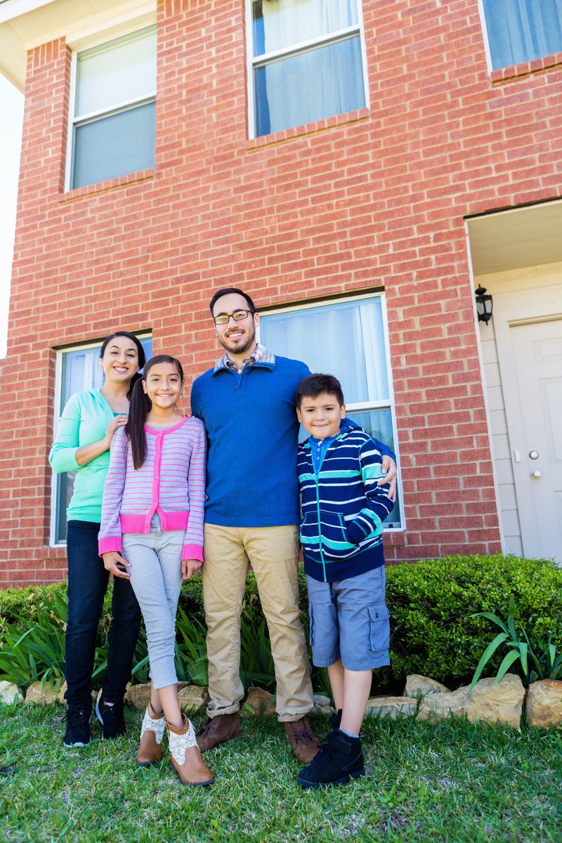 Mid adult Hispanic couple and their two children stand in front of their new home. They have a pre teen daughter and an elementary age son. They are standing in front of the bushes in the front yard and the red brick two story house is behind them.