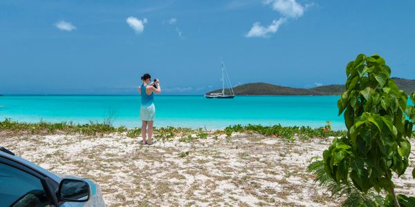 A woman takes a photo of a sailboat on turquoise waters from a sandy beach.