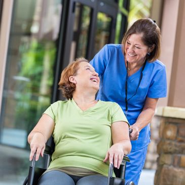 Caregiver smiles while pushing a woman in a wheelchair outside.
