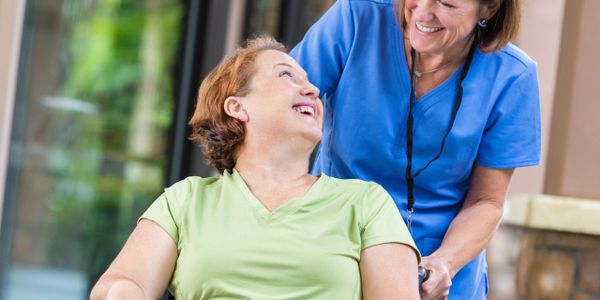 Caregiver in blue scrubs pushes smiling elderly woman in a wheelchair outdoors.