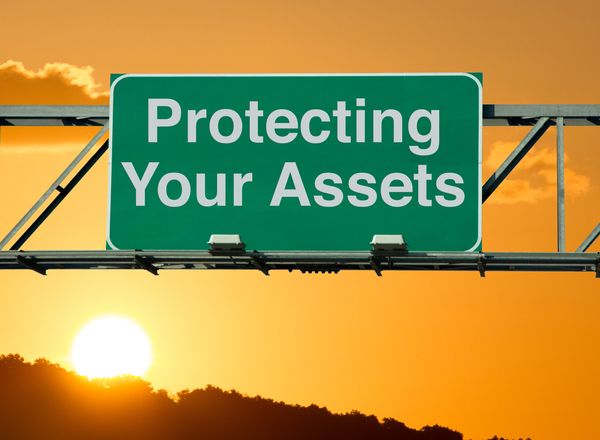Green highway sign stating Protecting Your Assets with the sun, a cloud, mountain, and a golden sky