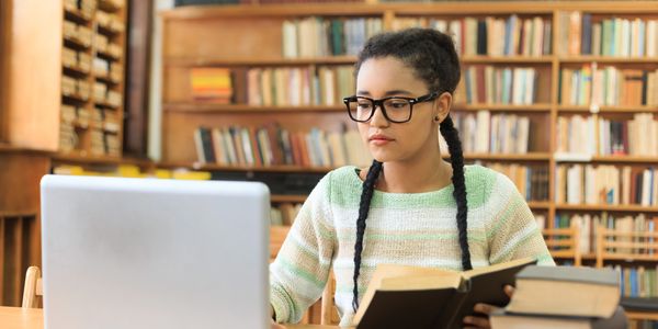 unna chica con un libro en las manos y un ordenador en frente estudiando en una biblioteca