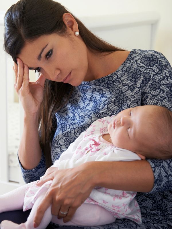 Tired mother holds sleeping baby, looking worried.