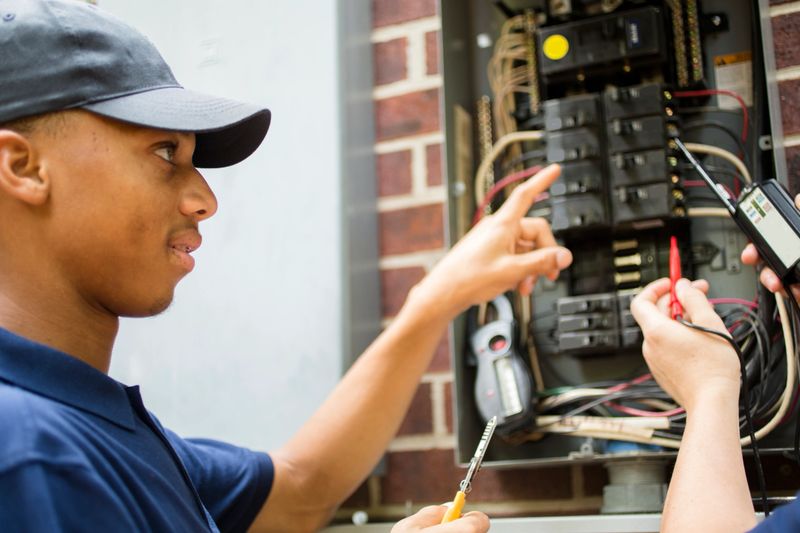 Two male electricians working on a residential breaker box.  The multi-ethnic team use testing tools and wear matching blue uniforms.  They are discussing next steps in the job repair project as they work.  Electric meter to side on brick home.