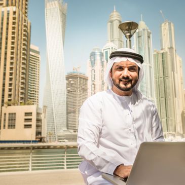 Middle Eastern man in traditional attire using a laptop outdoors in a modern city.