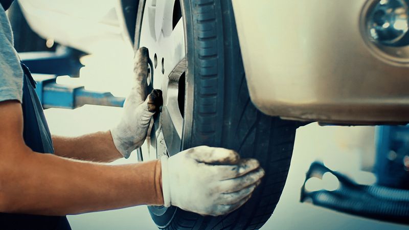 Closeup of unrecognizable male mechanic replacing car tire on front right hand wheel at a workshop. He's using pneumatic screwdriver while the car is lifted on a car jack. Side view.