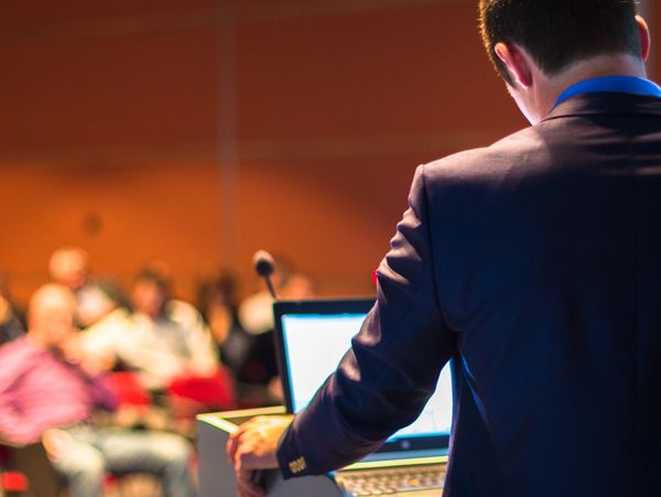 Man in suit giving a presentation to an audience with a laptop and microphone.
