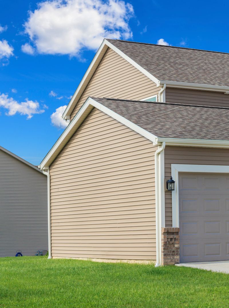 A new home with tan colored vinyl siding. Green grass and blue sky with clouds is in the photograph.