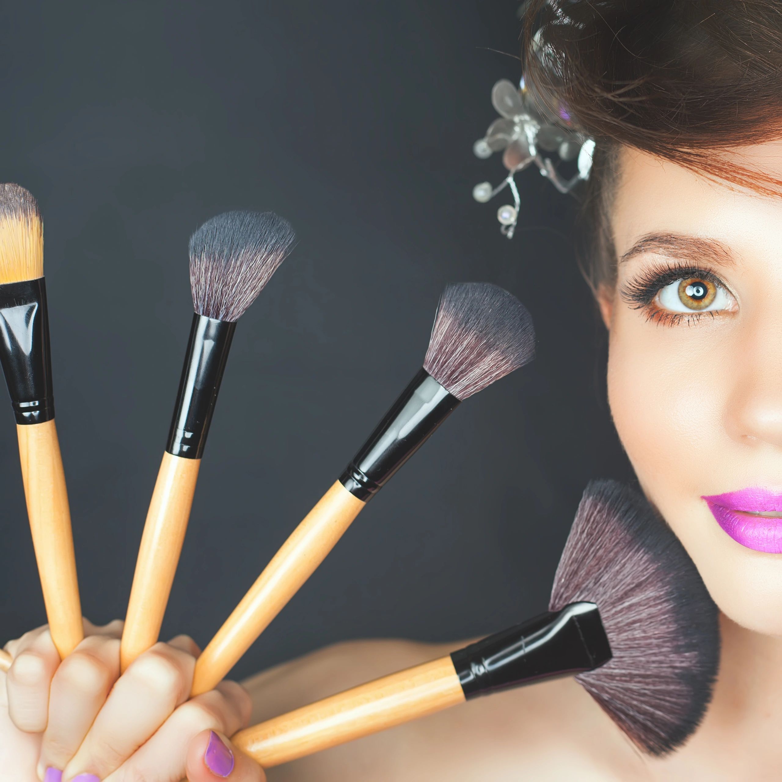 Close-up of a woman holding makeup brushes with purple lipstick and eye makeup.