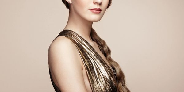 Elegant woman with long braided hair and metallic gold dress against beige background.