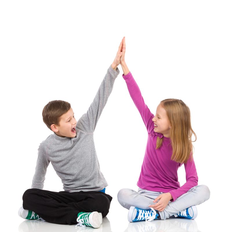Happy boy and girl sitting on the floor with legs crossed and giving high-five to each other. Full length studio shot isolated on white.