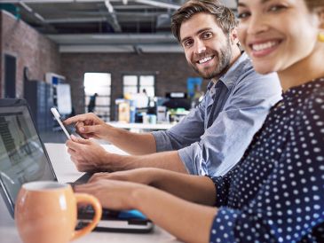 Two coworkers smiling and working together on laptops in a modern office.