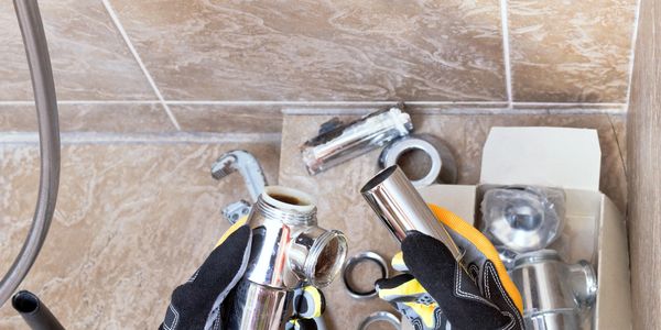 Hands in gloves holding chrome plumbing pipes with tools on tiled floor.