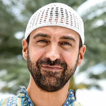 Man with a beard wearing a white knit cap and patterned shirt outdoors.