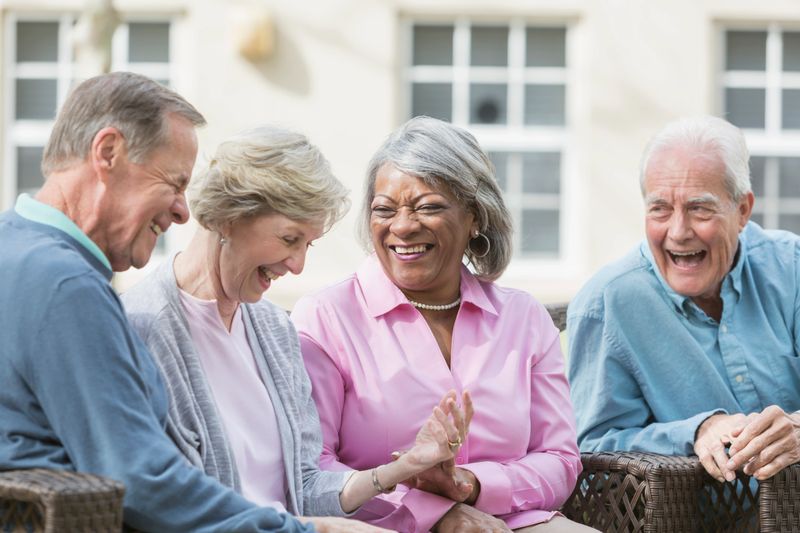 A group of four multi-ethnic seniors sitting together on patio furniture outdoors, talking and laughing. They are in retirement, relaxed and enjoying spending time with friends.