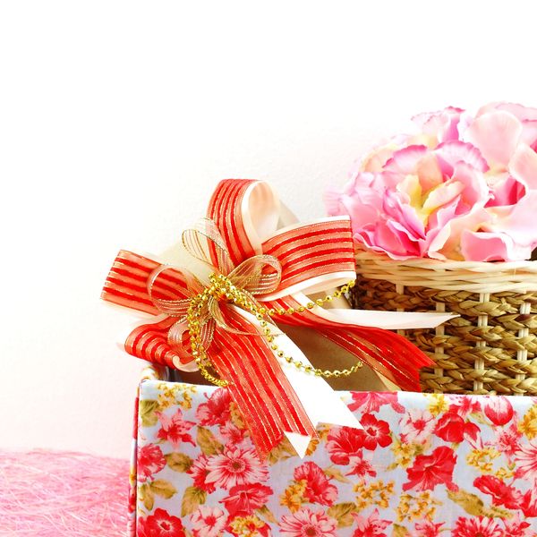 Gift box with a red and gold ribbon next to a basket of pink flowers.