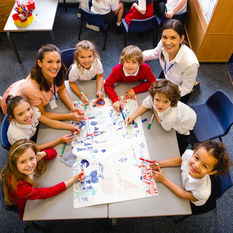 A medium group of children are sitting around a desk with two teacher and drawing pictures and brainstorming on a large sheet of paper. The image has been shot from above and the children and students are looking up smiling at the camera.