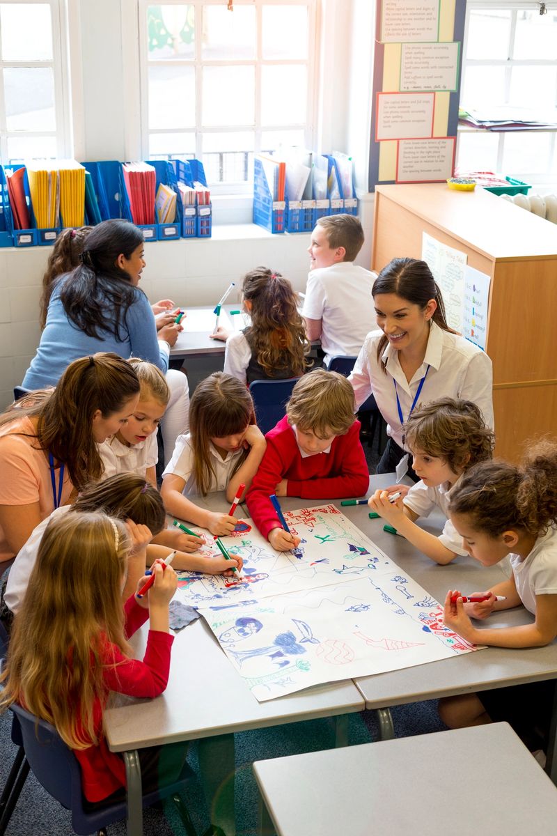 A large group of children are sitting around two desks in a classroom with teachers supervising. They are being creative drawing pictures and brainstorming on a large sheet of paper. The image has been shot from above and the children and students look happy.