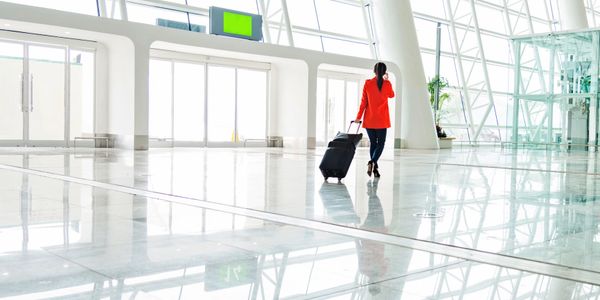 A woman in a red jacket walking with a suitcase in a bright airport terminal.