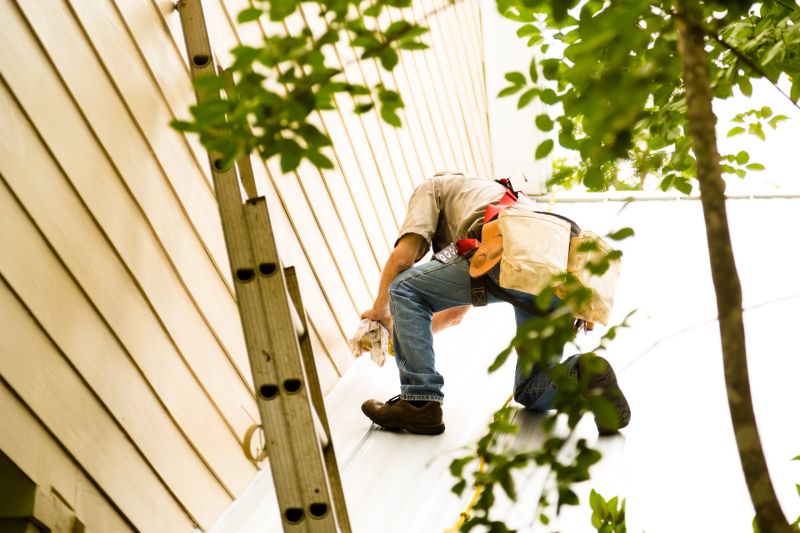 Repairman roofer repairs a tin, metal, or aluminum roof on a home. He has a ladder and tools in tool belt and wears a safety harness.  Construction, blue collar worker. Outdoors.