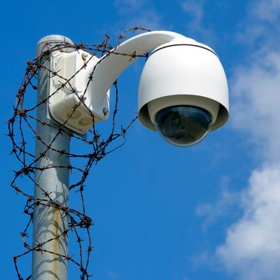 Security camera wrapped with rusty barbed wire on a pole against blue sky.