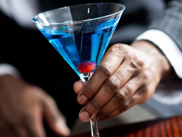 Hand holding a blue cocktail in a martini glass with a cherry inside.