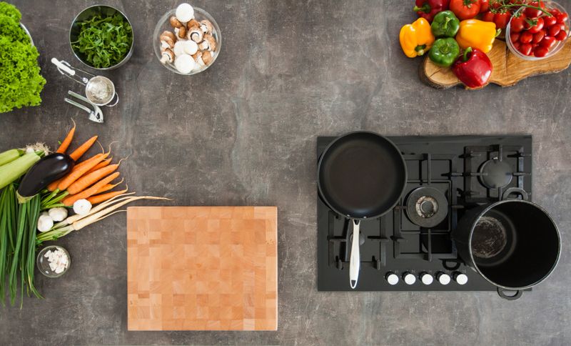 Overhead view of kitchen counter, stove with empty cooking and frying pan, vegetables and various cooking ingredients.
