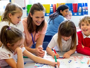 Teacher happily watches children drawing and coloring together in classroom.