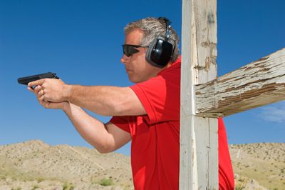 Man in red shirt aiming a handgun at an outdoor shooting range.