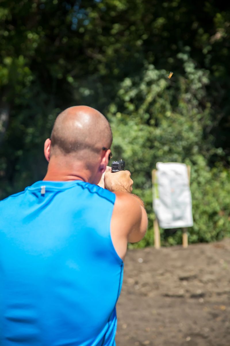 Over the shoulder view of a man shooting a handgun at a homemade outdoor target. Gun powder and the ammo casing can be seen flying in the air, as the gun has just been fired.
