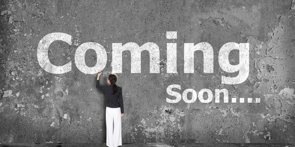 Woman standing at dark gray wall writing "Coming Soon" in white chalk.
