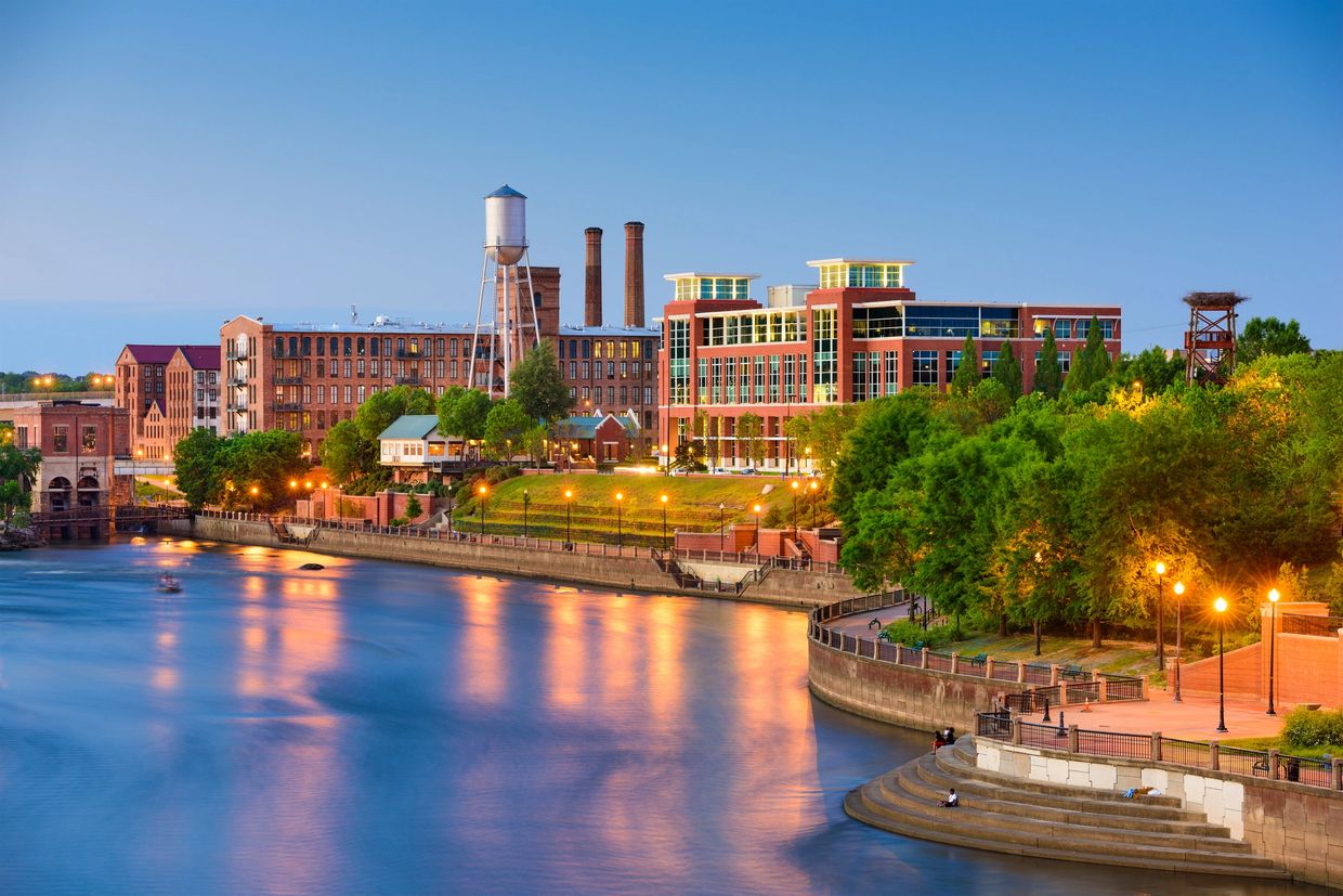A serene riverside cityscape at dusk with illuminated buildings and streetlights reflecting on the water.