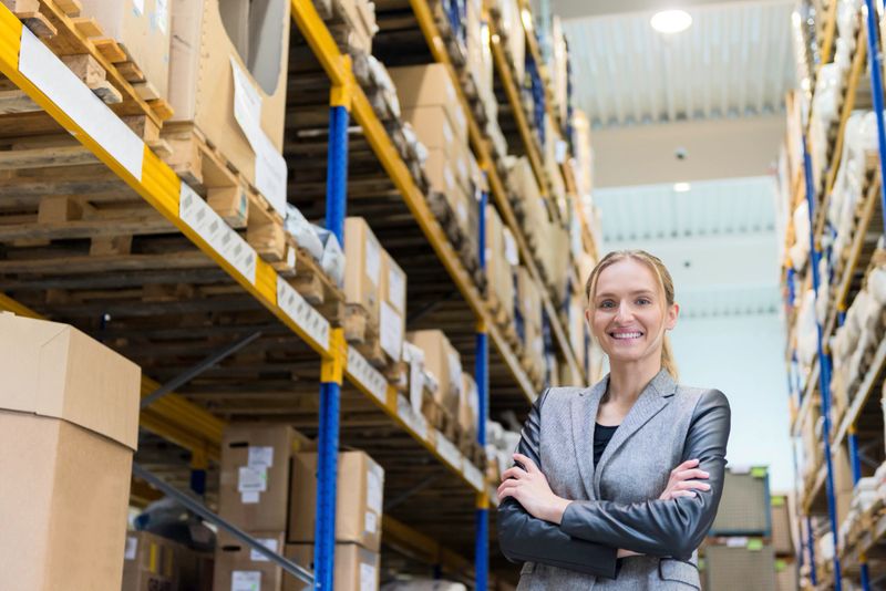 Low angle view portrait of a confident young woman supervisor with arms crossed in warehouse. Young blond woman standing at distribution warehouse and wearing elegant suit. Industrial boss sincerely smiling and looking at camera. Logistic worker working in a large distribution warehouse. Large distribution storage in background with racks and shelfs full of packages, boxes, pallets and crates ready to be delivered. Logistics, freight, shipping, receiving.