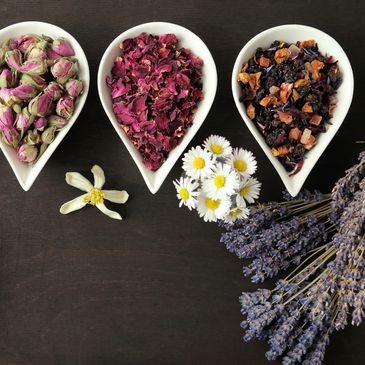 Four white spoons with different dried flowers and herbs on dark wood, surrounded by fresh flowers.