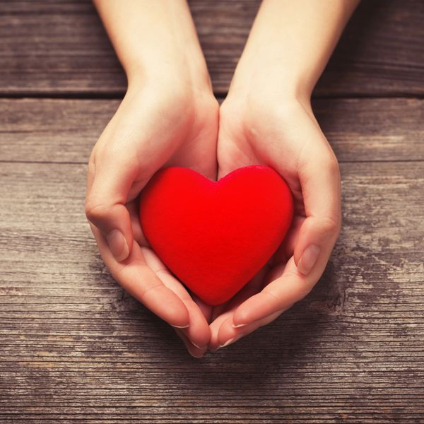 Hands holding a red heart on wooden surface.