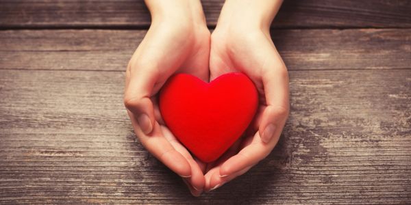 Hands gently holding a red heart on a wooden surface.