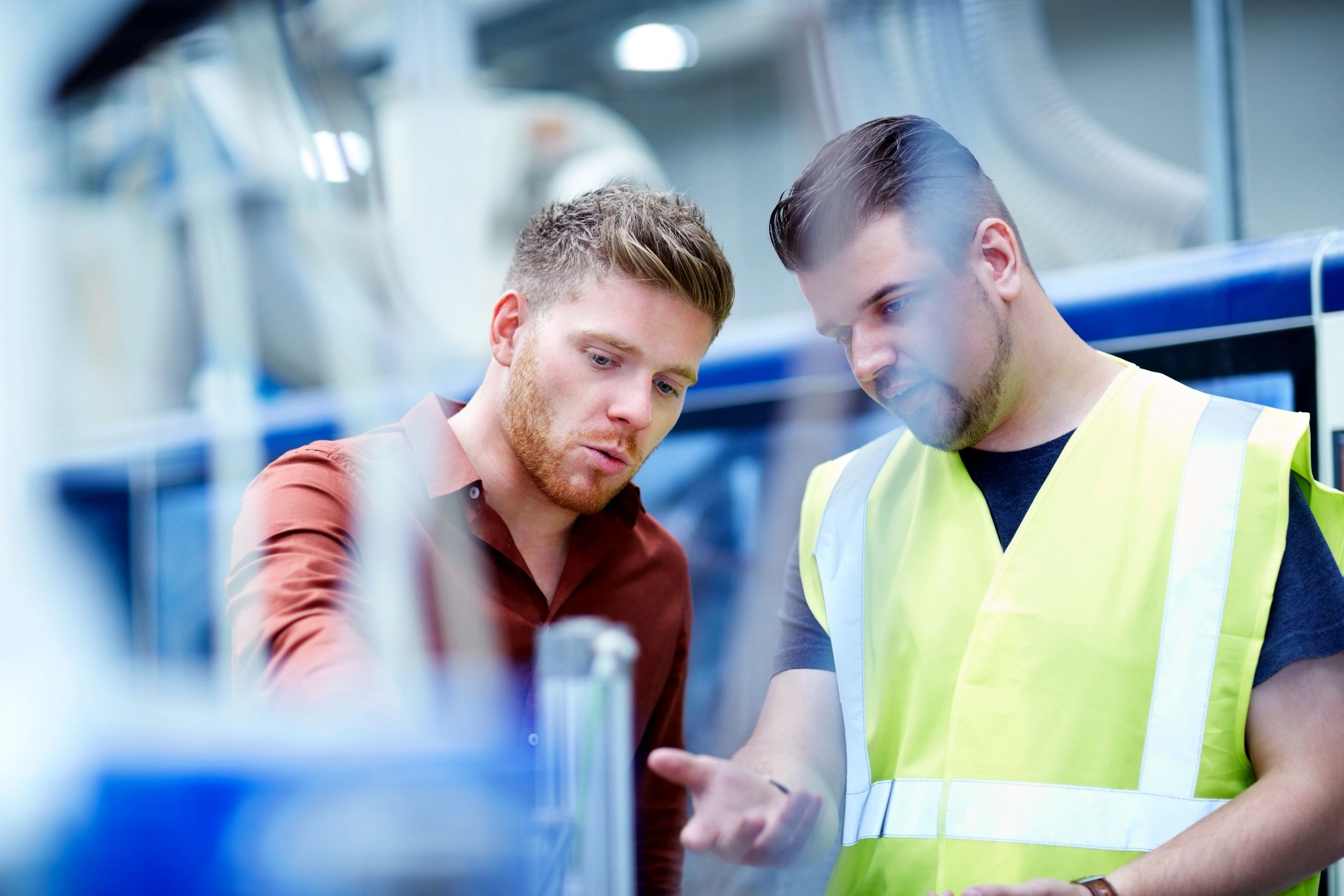 Two men examining something intently, one wears a safety vest.