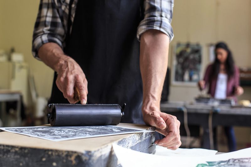 Young woman and man working at workshop. Close up of male lithograph using a printing roller. Focus on foreground.