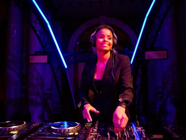 A female DJ smiling while mixing music with headphones in a vibrant club setting.