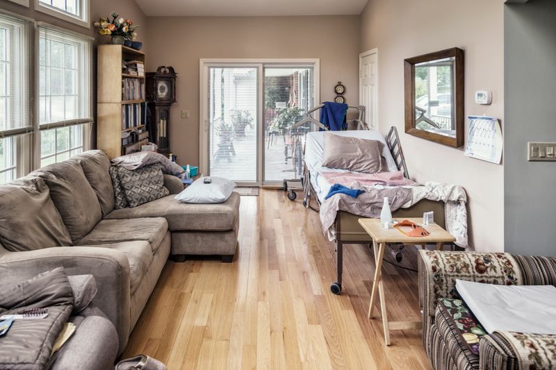 Horizontal lengthwise view of a long, narrow, bright, but very cluttered residential home living room with a rolling, wheeled, tilting, adjustable height hospital bed and disabled patients medical hoist equipment as the temporary main features.
