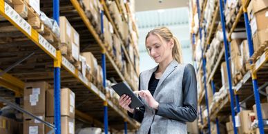 A woman using a tablet inside a warehouse with tall shelves of boxes.