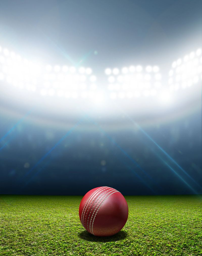 A cricket stadium with a red leather cricket ball on an unmarked green grass pitch at night under illuminated floodlights