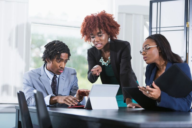 A team of three African American businesspeople working together on a project. The young woman standing in the middle with her hand on her hip is in charge, telling the young man sitting at the table what to do. He is typing on his digital tablet as she points to the screen. Another woman sitting at the table is taking notes.