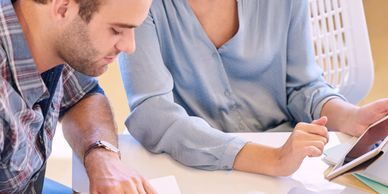 Two colleagues collaborating over notes and a tablet at a desk with a laptop.