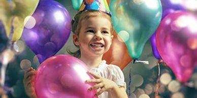 Smiling child wearing party hat surrounded by colorful balloons.