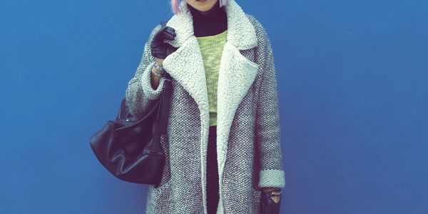 Person in a cozy gray coat with black gloves and a black bag against a blue background.