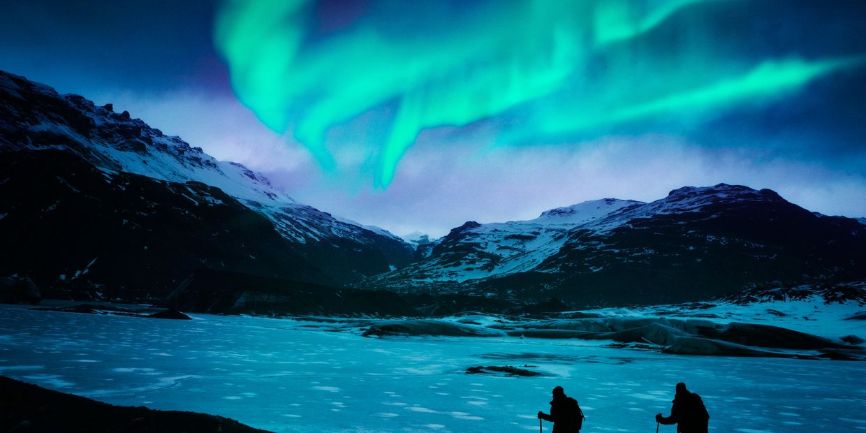 Two hikers under the Northern Lights in a snowy mountain landscape.
